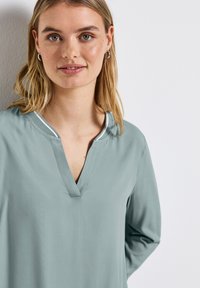 Blouse vert clair à manches longues avec un décolleté en V et un contour blanc contrastant autour du col. Texture de tissu lisse et douce.