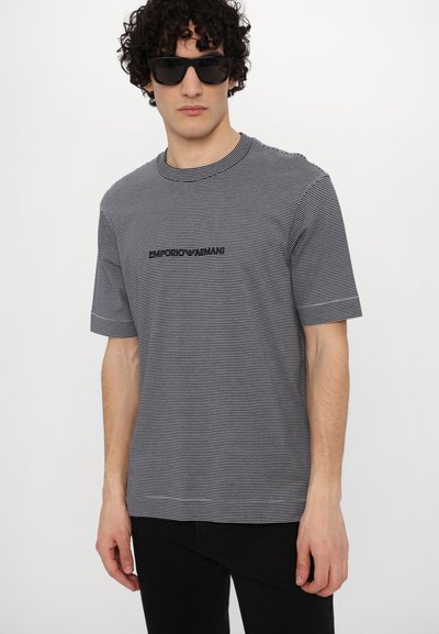 T-shirt às riscas, de manga curta, em algodão cinza e preto, com decote redondo e um logótipo bordado na frente. Acompanhado por óculos de sol.