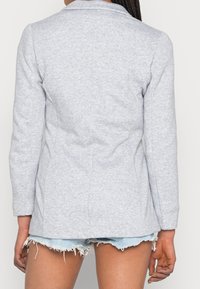 Blazer gris clair en tissu doux, présentant une coupe ajustée, des manches longues, des détails de couture subtils et un col à revers cranté.