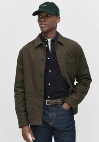 Olivengrøn jakke med knapper, en brystlomme og krave, lagdelt over en marineblå skjorte. Båret med mørk denim og et vævet bælte.