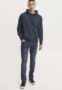 Sudadera azul marino con letras doradas, bolsillo frontal y cordones. Combinada con jeans azules y zapatillas negras, de pie contra un fondo claro.