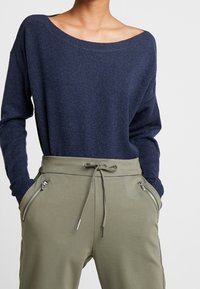 Pull en maille bleu marine avec un design épaules dénudées, associé à un pantalon de jogging vert olive doté d'un cordon de serrage et de poches zippées.