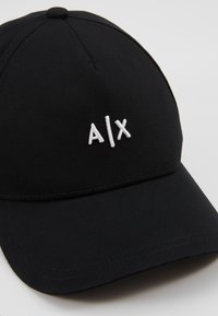 Gorra de béisbol negra hecha de tela, con visera curva y logo bordado en blanco "A/X" en la parte frontal. Textura suave, diseño simple.