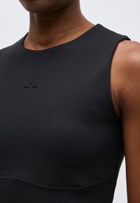 Κοντινό πλάνο ενός ατόμου που φοράει μαύρο, ribbed τοπ χωρίς μανίκια με ένα μικρό, διακριτικό λογότυπο κοντά στη γραμμή του λαιμού.