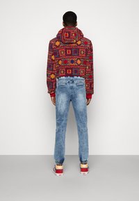 Hoodie s červeným a žlutým vzorem a zkráceným střihem, kombinovaný se světlými modrými džíny a mnohobarevnými teniskami Vans.