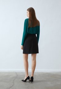 Jupe noire au design ajusté, associée à un blouse à manches longues couleur sarcelle. Le mannequin porte des chaussures à talons hauts noires, avec des cheveux lisses visibles.