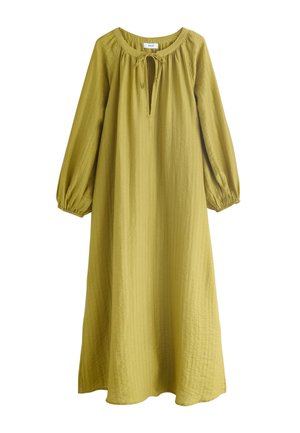 Femme aux longs cheveux noirs debout sur des rochers, portant une robe ample à manches longues de couleur vert olive avec une fente à l'avant et des sandales noires.
