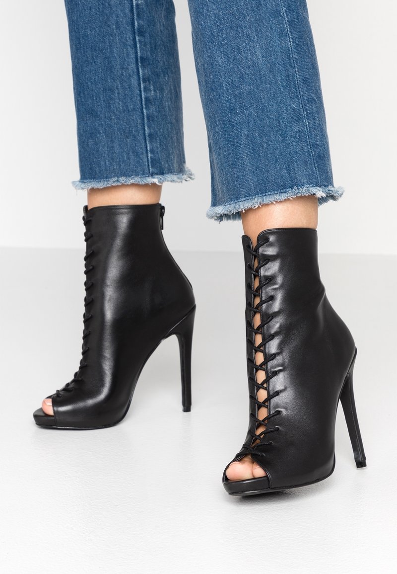 Bottines à talons en cuir noir avec bout ouvert et lacets. Présentent une texture lisse et un design élégant et ajusté.