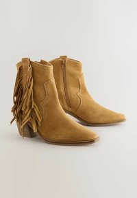 Botas de tobillo de gamuza marrón con detalles de flecos, punta afilada, cremallera lateral y tacón apilado. Textura suave y acentos de costura decorativa.