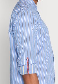 Blåstribet skjorte med knapper, der har lodrette hvide striber, en opbukket ærme med detaljer i flere farver og en klassisk krave.