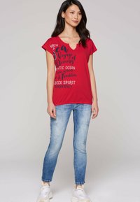Red short-sleeve t-shirt featuring a V-neck and graphic text design. Paired with light blue skinny jeans and white sneakers.