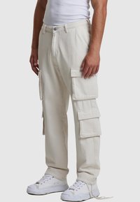 Pantalones cargo beige hechos de algodón, con múltiples bolsillos laterales, un ajuste relajado y detalles de cuerda ajustables en los tobillos. Llevados con zapatillas blancas.