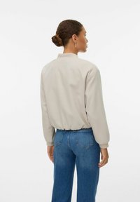 Lichtbeige bomberjack met een kraag, elastische tailleband en lange mouwen, gecombineerd met blauwe highwaisted jeans.