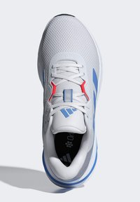 Chaussure de sport blanche avec une tige en mesh texturé, des accents rouges et bleus, des lacets plats et une semelle rembourrée. Marquage notable sur le bout.