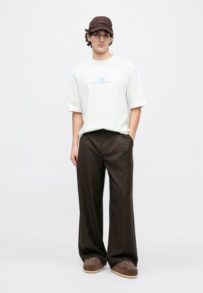 Tricou alb cu mâneci scurte și logo albastru, pantaloni maro cu picioare largi, șapcă maro și pantofi maro slip-on cu un model unic.