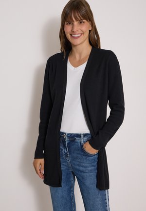Vrouw glimlacht, draagt een zwarte open cardigan, wit t-shirt en blauwe spijkerbroek met haar rechterhand in haar zak tegen een effen achtergrond.