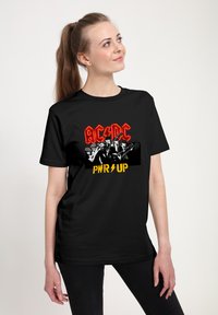 Czarny bawełniany t-shirt z żywą czerwoną i żółtą grafiką z napisem "AC/DC" oraz "PWR UP" obok członków zespołu trzymających instrumenty.