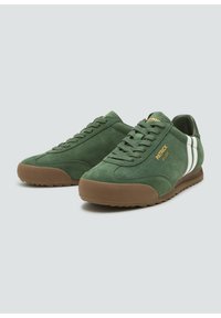 Zielone zamszowe trampki z brązowymi gumowymi podeszwami, z białymi paskami po bokach i złotym logo. Klasyczny sportowy design z sznurówkami z przodu.