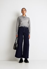 Siv pulover iz pletene tkanine s ovratnikom, v kombinaciji s širokimi temno modrimi jeans hlačami. Model drži črno torbico in nosi temne čevlje.