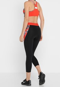 Reggiseno sportivo rosso con un motivo floreale, abbinato a leggings neri a tre quarti con una vita prominente. Scarpe da ginnastica nere completano il look.
