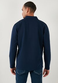 Camisa polo de manga larga en color azul marino con cuello clásico, textura suave y corte recto. Se lleva con jeans azules, mostrando un diseño sencillo en la parte posterior.