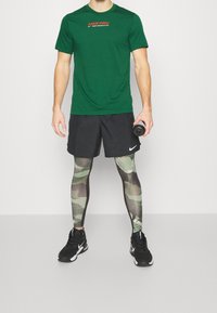 Hombre vistiendo una camiseta verde de Nike Pro, pantalones cortos negros de Nike, leggings de camuflaje y zapatillas negras de Nike, sosteniendo una botella de agua gris.