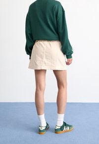 Groene sweatshirt, beige rok en groene sneakers met witte accenten. Het materiaal lijkt zacht; de sweatshirt heeft een losse pasvorm, de rok is eenvoudig en casual.