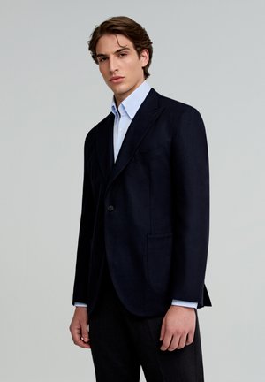 Granatowy wełniany żakiet z wykładanymi klapami, zapięciem na jeden guzik i dwiema przednimi kieszeniami, połączony z jasnoniebieską koszulą.