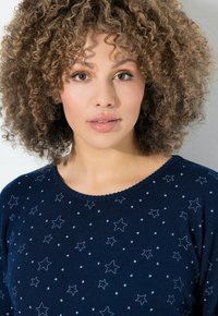 Frau mit lockigem Haar, die einen dunkelblauen Pullover mit weißen Sternmustern trägt und direkt in die Kamera schaut, vor einem schlichten Hintergrund.