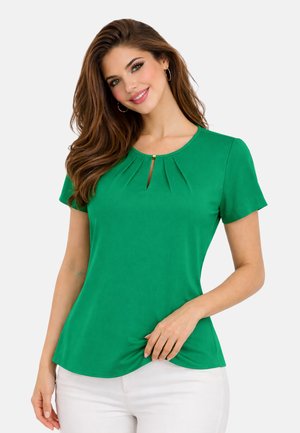 Kobieta uśmiechająca się, ubrana w zieloną bluzkę z krótkim rękawem z plisami z przodu i detalem z wycięciem, w połączeniu z białymi spodniami, na jednolitym tle.