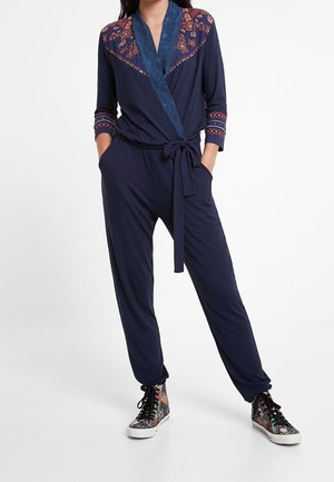 Woman wearing navy blue jumpsuit with floral embroidery on shoulders and cuffs, tied waist, and multicolor high-top sneakers.