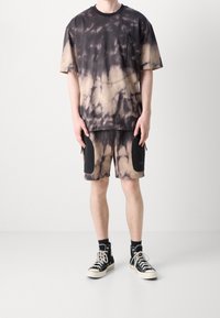Camiseta de tie-dye en negro y beige con un bolsillo frontal con cremallera, combinada con pantalones cortos que tienen bolsillos laterales negros y cremalleras. Llevado con zapatillas negras.