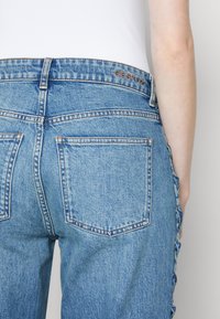 Světle modré džíny z denimu s vysokým pasem, zadními kapsami a knoflíkovým detailem podél strany. Logo "GESTUZ" je vyraženo na pásu.