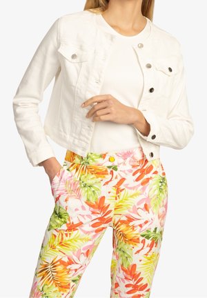 Femme portant une veste courte blanche et un pantalon à motifs floraux avec des feuilles orange, jaune, vertes et roses, une main dans la poche.