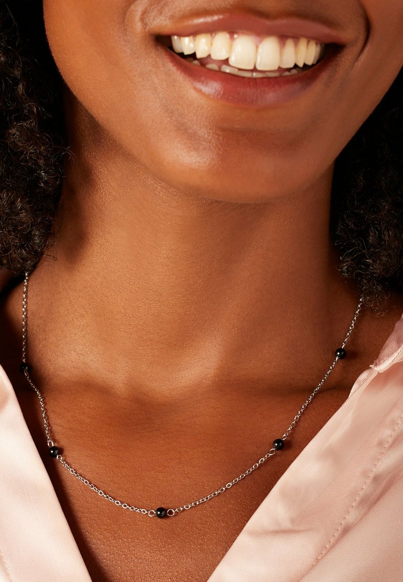 Zilveren ketting met zwarte kralen, delicate schakels en een elegant ontwerp, gedragen om een nek met een gladde huid en een roze kledingstuk.