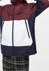 Kleurgeblokte jas met een bordeauxrode bovenkant, een wit middenstuk en een marineblauwe onderkant. Voorzien van ritsen en een capuchon. Zacht, lichtgewicht materiaal.