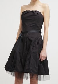 Robe noire sans bretelles avec un corsage ajusté et une jupe évasée à volants. Présente un tissu texturé, une ceinture en satin avec un nœud et une couche en tulle en dessous.