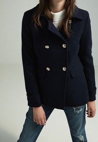 Marine uld dobbeltbrystet frakke med guldknapper, skræddersyet pasform og bred krave. Båret over en lys struktureret sweater og slidte jeans.