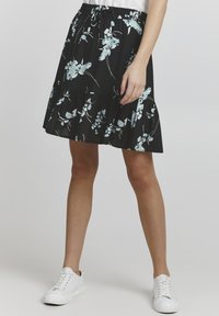 Fustă neagră cu flori, având modele albastre deschise, volane în straturi și talie elastică, purtată cu adidași albi. Textura materialului este netedă.