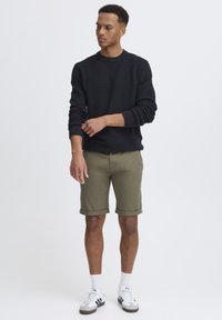Pull en maille côtelée marine et shorts vert olive, avec des manches retroussées, associés à des chaussettes blanches et des baskets noir/blanc, se tenant devant un fond uni.