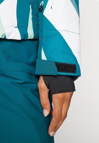 Sinise ja valge geomeetrilise mustriga kanal-jakk. Omab takjakinnitustega mansette ja musta sisemise varruka. Vooderdus on tikandiga ja sileda viimistlusega.