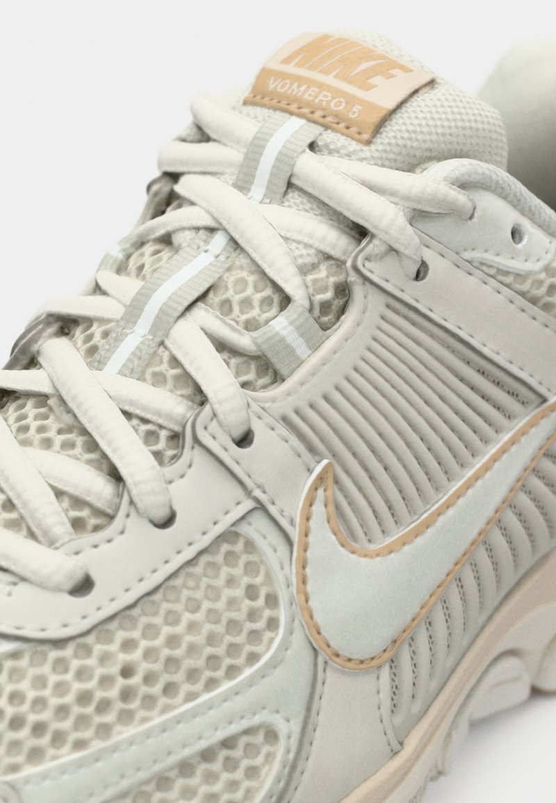 Helle graue Nike Vomero 5 Sneaker mit Mesh-Obermaterial, gepolsterter Ferse und beigen Akzenten. Verfügt über eine Gummisohle und strukturierte Overlays.