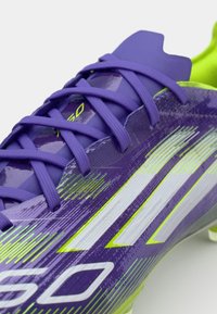 Purple soccer cleat with vibrant yellow accents, textured synthetic upper, and contrasting white stripes. Features thin laces and molded studs.