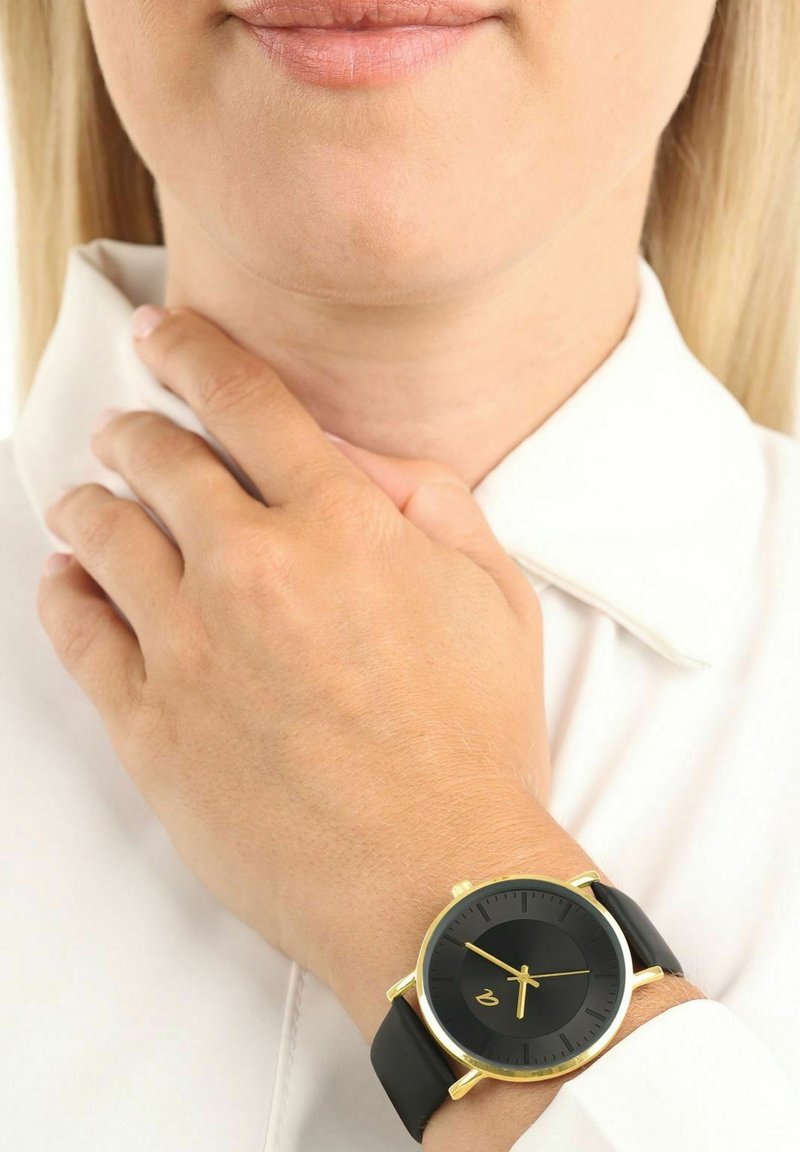 Close-up van een persoon die een zwart-gouden horloge met een zwarte wijzerplaat draagt, terwijl hij met zijn linkерп hand zijn nek vasthoudt.