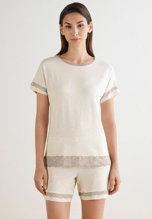 Krämfärgad loungewear-set som består av en kortärmad topp och shorts, accentuerad med delikat spetskant vid ärmarna och fållen.