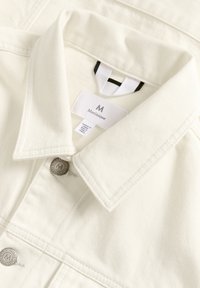 Jachetă din denim albă cu guler ascuțit, închidere cu buton argintiu și detalii cusute. Prezintă un etichetă cu numele brandului "Matinique" vizibilă.