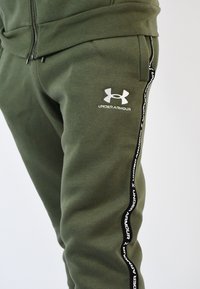Pantalones jogger verde oliva, hechos de material suave, con un logotipo blanco de Under Armour y franjas laterales negras con texto repetido. Cintura ajustable.
