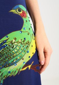 Marinblå klänning med en grön fågodesign prydd med paljetter i blått och guld, kompletterad med ett texturerat tyg och intrikata detaljer.