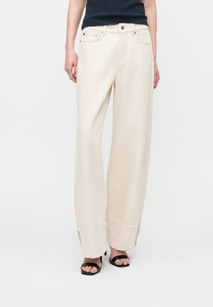 Kobieta ubrana w kremowe spodnie z wysokim stanem i szerokimi nogawkami z postrzępionymi brzegami oraz bocznymi rozcięciami, w zestawieniu z czarnymi sandałami na obcasie z odkrytymi palcami i czarnym, bezrękawnym topem.