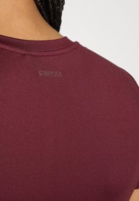 Sportsku majicu u boji burgundac s okruglim izrezom i kratkim rukavima, s diskretnim reljefnim logotipom "STRONGER" na leđima ispod ovratnika.